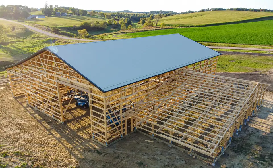 A post-frame building under construction with its steel roof on and wall framing exposed, farm fields behind